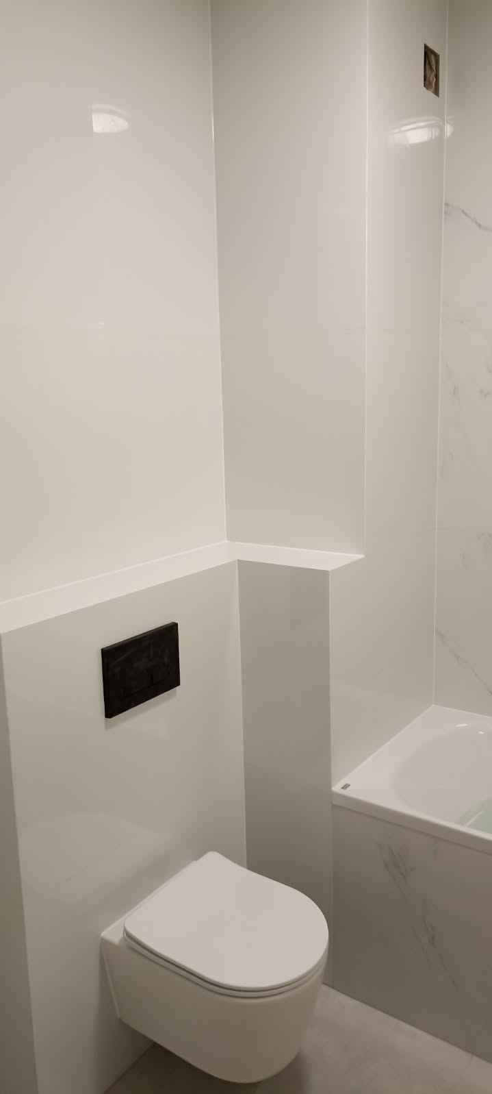 Nowoczesna łazienka z białą armaturą i panelami ściennymi, widoczna wisząca toaleta z ciemnym przyciskiem spłukującym i fragment wanny obłożonej płytkami imitującymi marmur.