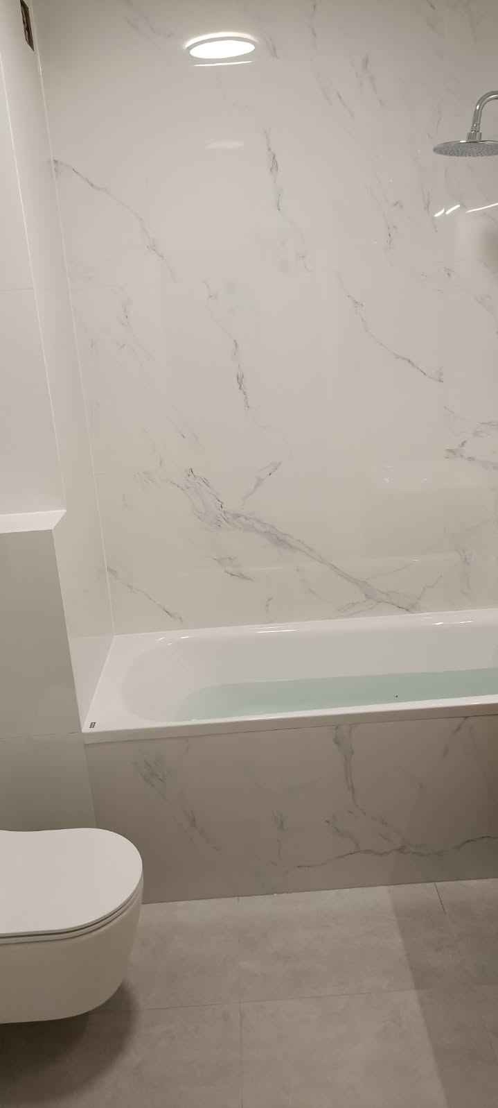 Nowoczesna łazienka z wanną wypełnioną wodą, białe płytki imitujące marmur na ścianach i szare płytki podłogowe, wisząca toaleta.