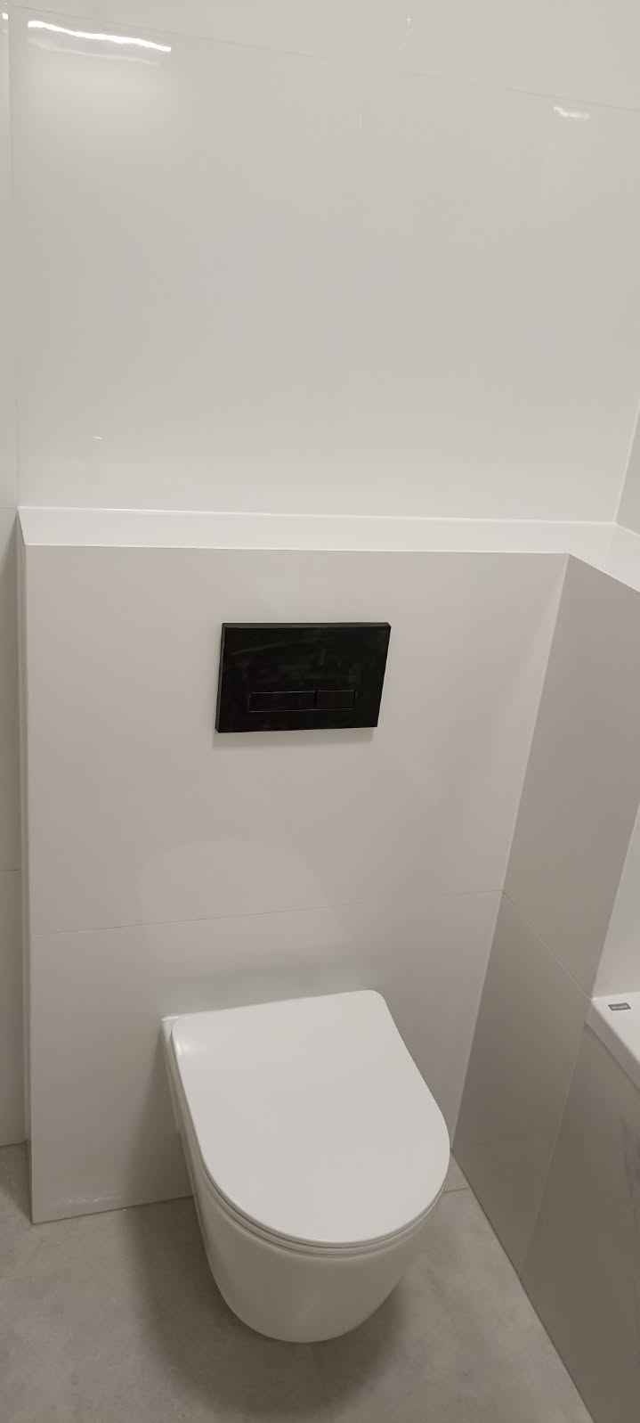 Świeżo wykończona, minimalistyczna łazienka z podwieszaną toaletą i czarnym panelem spłukującym, wykończona białymi, błyszczącymi płytkami.