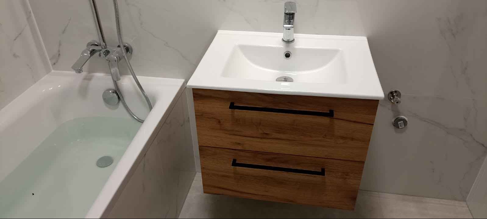 Nowoczesna łazienka z białą wanną wypełnioną wodą i umywalką nablatową z szafką z drewnopodobnym fornirem i czarnymi uchwytami, na tle marmurowych płytek.