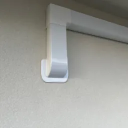 Biały, prostokątny kanał wentylacyjny przymocowany do szarej, teksturowanej ściany zewnętrznej budynku, widoczne mocowanie kanału do ściany.