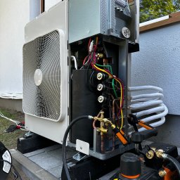 ECO CLIMA GAS - Krzysztof Murawski CALUNA - Otwarta obudowa pompy ciepła podczas instalacji na zewnątrz budynku. Widoczne rury, kable, elementy montażowe i narzędzia.