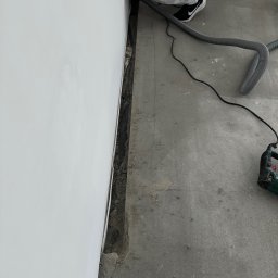 ECO CLIMA GAS - Krzysztof Murawski CALUNA - Instalator w trakcie wykonywania bruzdy w ścianie przy podłodze, używa odkurzacza przemysłowego i narzędzi do cięcia. Widoczny przewód instalacyjny.