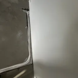 Białe rury instalacyjne ułożone przy ścianie, częściowo w bruździe w szarej posadzce. Widoczne gniazdko elektryczne na ścianie.