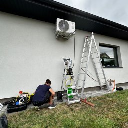 ECO CLIMA GAS - Krzysztof Murawski CALUNA - Instalacja klimatyzacji zewnętrznej na elewacji domu. Widoczny pracownik, drabina, narzędzia i butla z czynnikiem chłodniczym. Oświetlenie dzienne, pochmurne niebo.