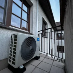 Biała jednostka zewnętrzna klimatyzacji Midea na balkonie z szarą podłogą, zamontowana na czarnych podkładkach antywibracyjnych. Widok na budynek przez balustradę.