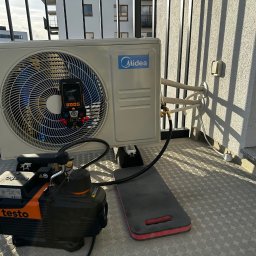 ECO CLIMA GAS - Krzysztof Murawski CALUNA - Biała jednostka zewnętrzna klimatyzacji Midea na balkonie, podłączona do pompy próżniowej Testo i miernika. Widoczne narzędzia i podkładka. Dobre oświetlenie.