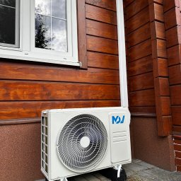 ECO CLIMA GAS - Krzysztof Murawski CALUNA - Biała jednostka zewnętrzna klimatyzacji zamontowana na brązowej, drewnianej elewacji domu z białym oknem. Widoczna rura odprowadzająca skropliny.