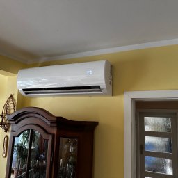 ECO CLIMA GAS - Krzysztof Murawski CALUNA - Biały klimatyzator ścienny z wyświetlaczem temperatury 27 stopni Celsjusza, zamontowany w pokoju z żółtymi ścianami, nad ciemnym kredensem.