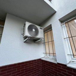 ECO CLIMA GAS - Krzysztof Murawski CALUNA - Biała jednostka klimatyzacji NOXA zamontowana na elewacji budynku z ceglaną podmurówką i oknem z kratami. Widok z dołu, światło dzienne.