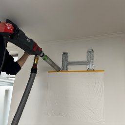 ECO CLIMA GAS - Krzysztof Murawski CALUNA - Wiercenie otworu w ścianie pod montaż klimatyzacji. Widoczne metalowe uchwyty, folia ochronna i wiertło z odsysaniem pyłu. Praca w trakcie wykonywania.