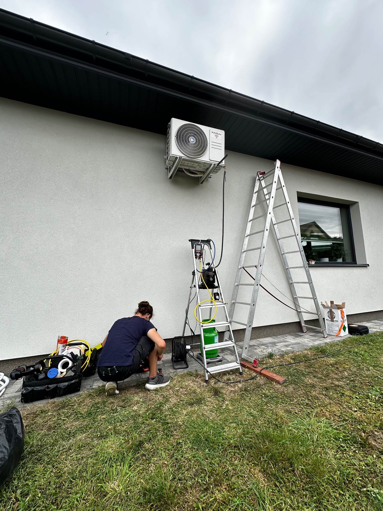 Serwisant na trawie przy ścianie domu, naprawia klimatyzator Kaisai. Widoczna drabina, narzędzia i butla z czynnikiem chłodniczym. Napełnianie układu chłodniczego.