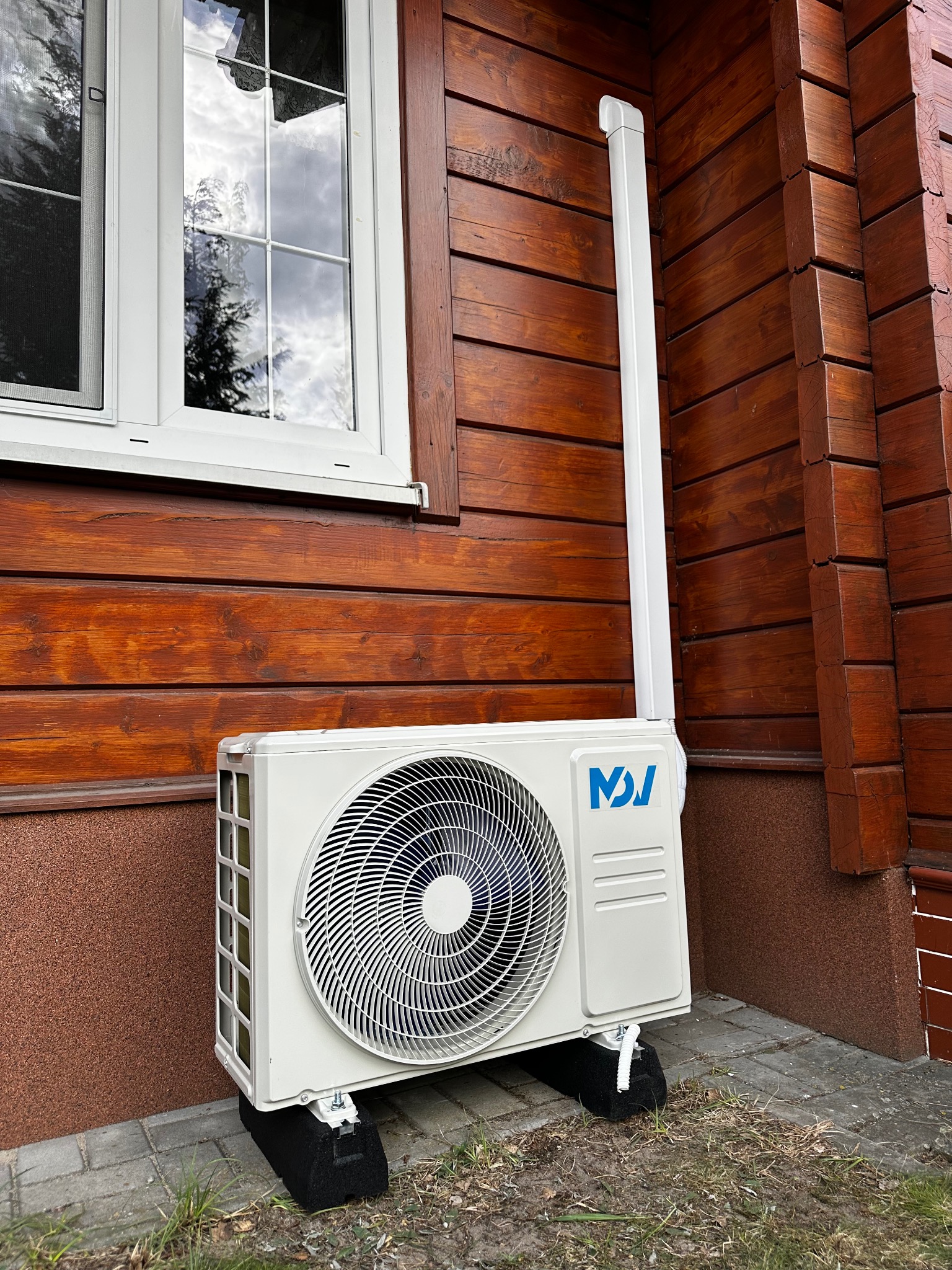 Biała jednostka zewnętrzna klimatyzacji zamontowana na brązowej, drewnianej elewacji domu z białym oknem. Widoczna rura odprowadzająca skropliny.