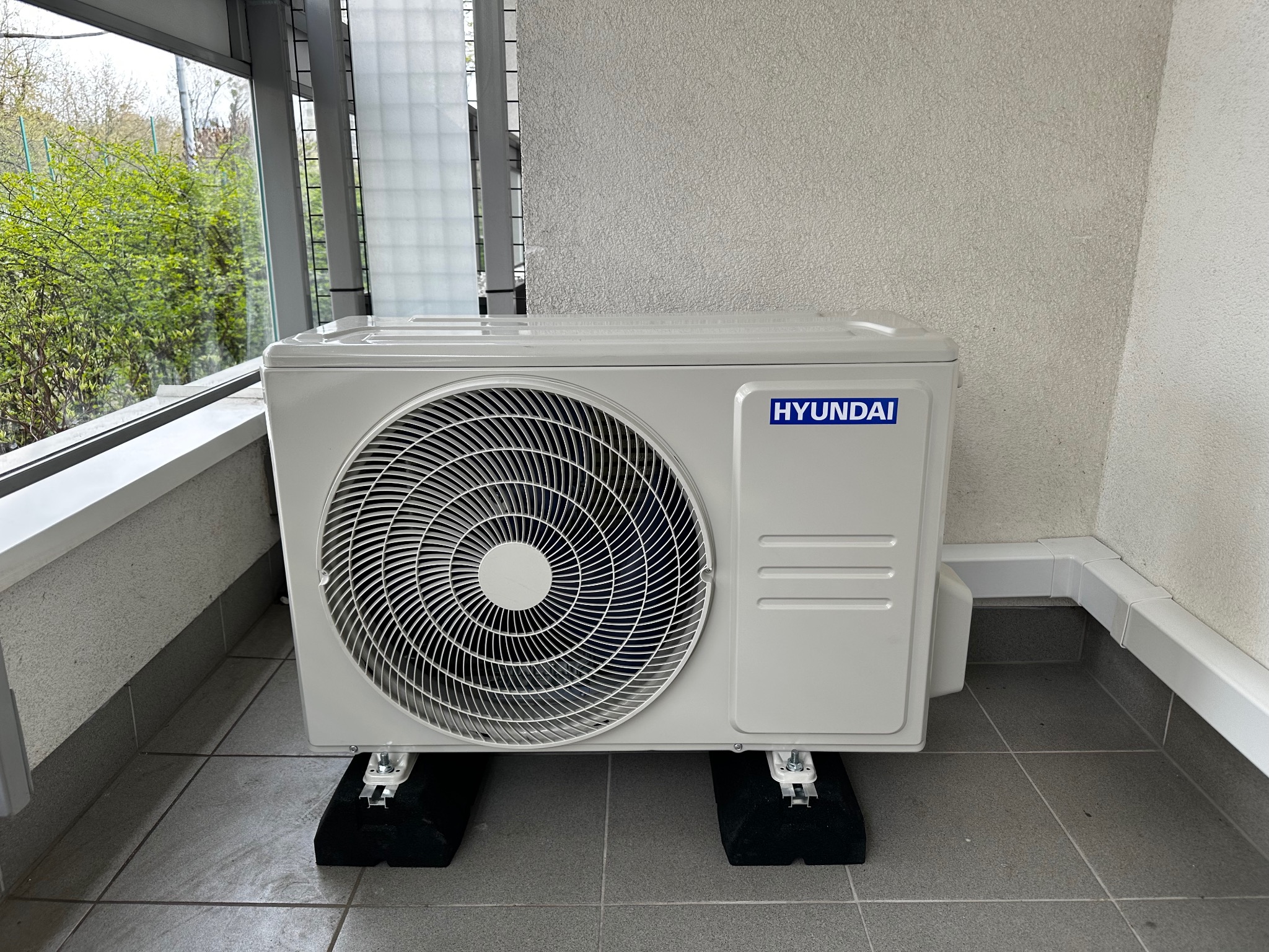 Biała jednostka zewnętrzna klimatyzacji Hyundai na balkonie, zamontowana na czarnych podkładkach antywibracyjnych. Widoczne szare płytki podłogowe i fragment ściany.
