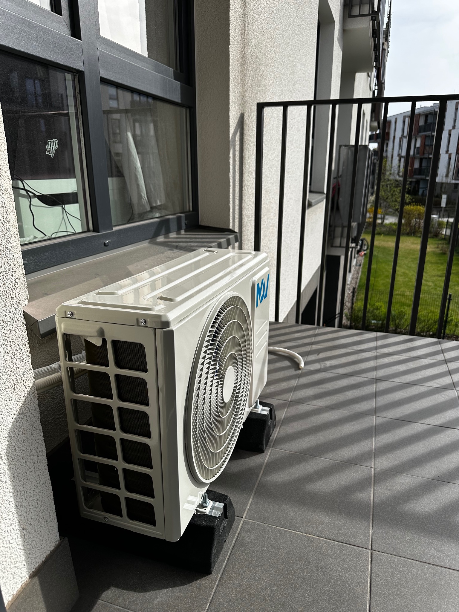 Biała jednostka zewnętrzna klimatyzacji na balkonie z szarymi płytkami, widok z boku, z fragmentem okna i balustrady w tle.
