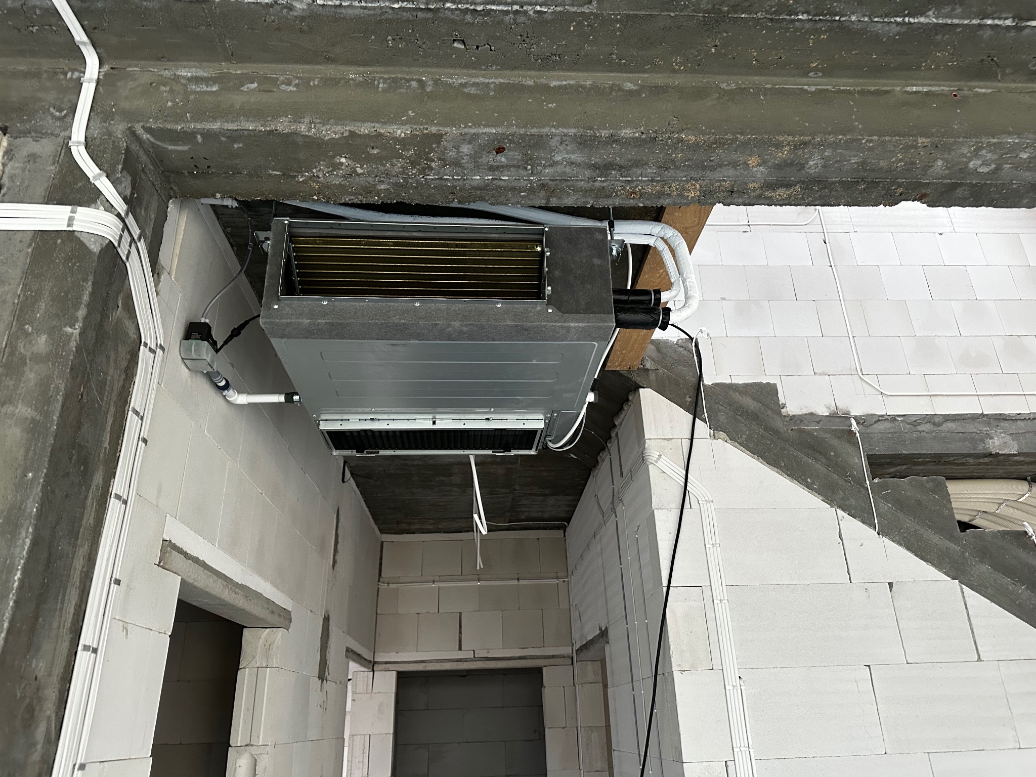 Sufitowa jednostka klimatyzacyjna zamontowana w surowym wnętrzu z białych bloczków, widoczne przewody i instalacje. Ujęcie z dołu, podkreślające etap montażu.