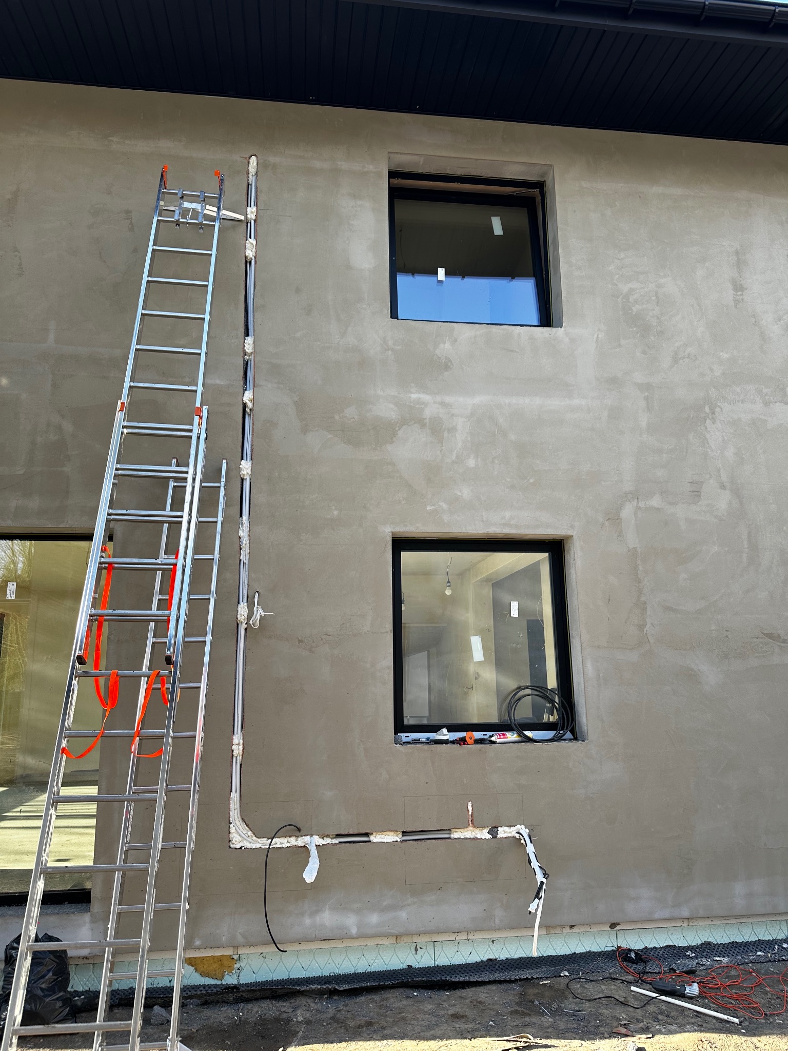 Elewacja budynku w trakcie prac instalacyjnych: rury prowadzone po ścianie, drabina oparta o mur, okna z czarnymi ramami. Widoczne materiały izolacyjne i narzędzia.