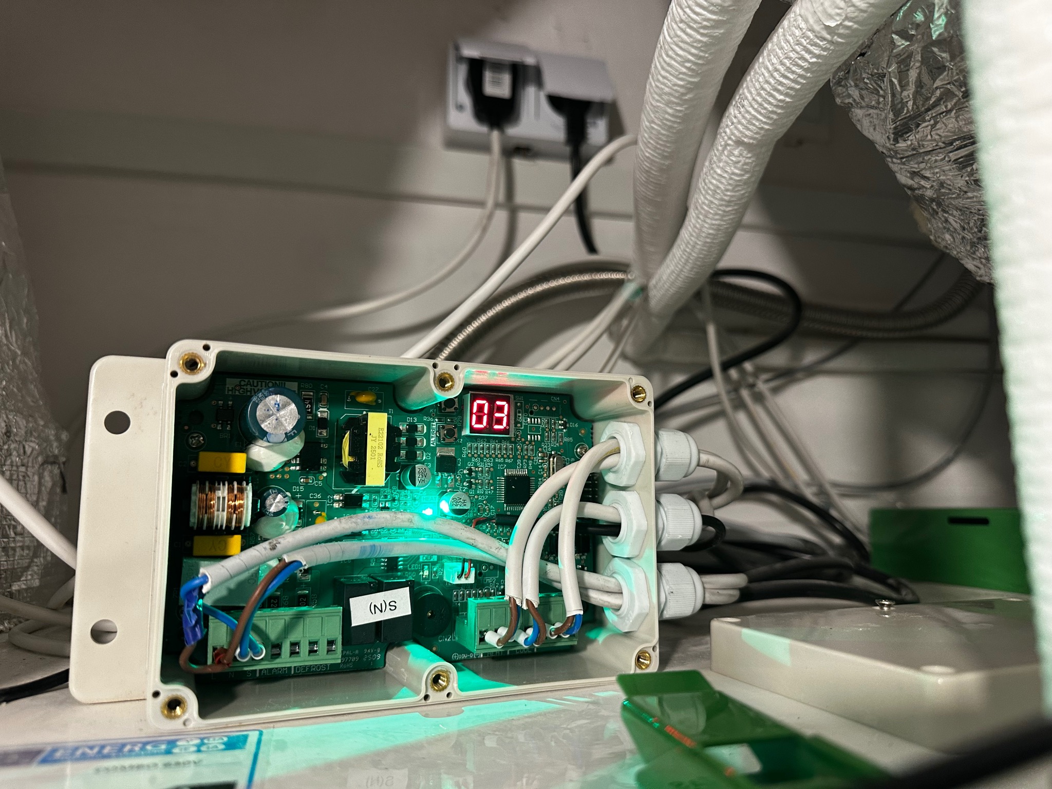 Otwarta skrzynka sterownicza z zieloną płytką drukowaną, wyświetlaczem LED i podłączonymi kablami, zamontowana w instalacji wentylacyjnej. Widoczne przewody i zasilanie.