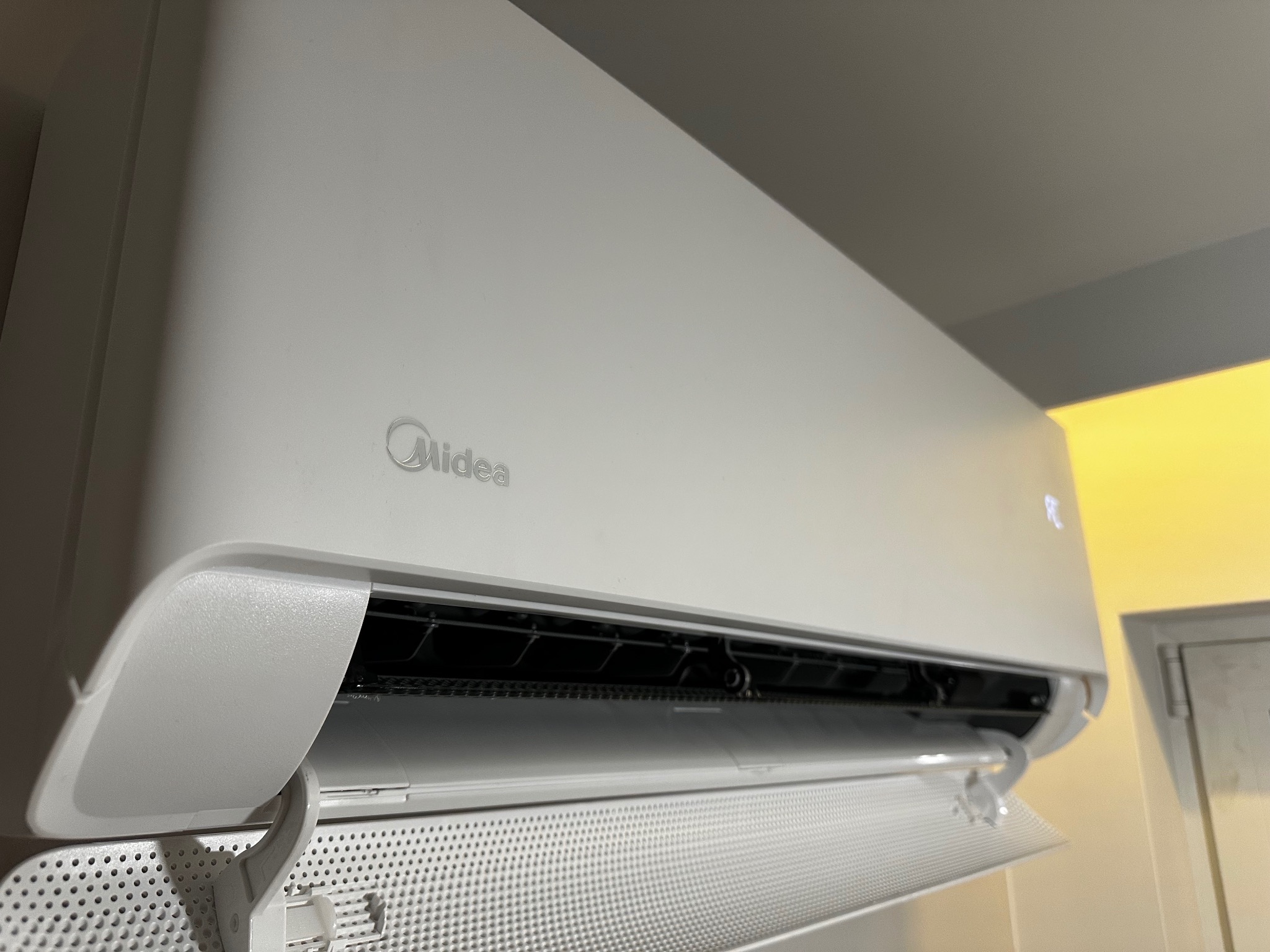 Biały klimatyzator Midea zamontowany na ścianie, widoczny z dołu pod kątem. Otwarta klapka dolna ukazuje perforowaną powierzchnię. W tle fragment ściany i drzwi.