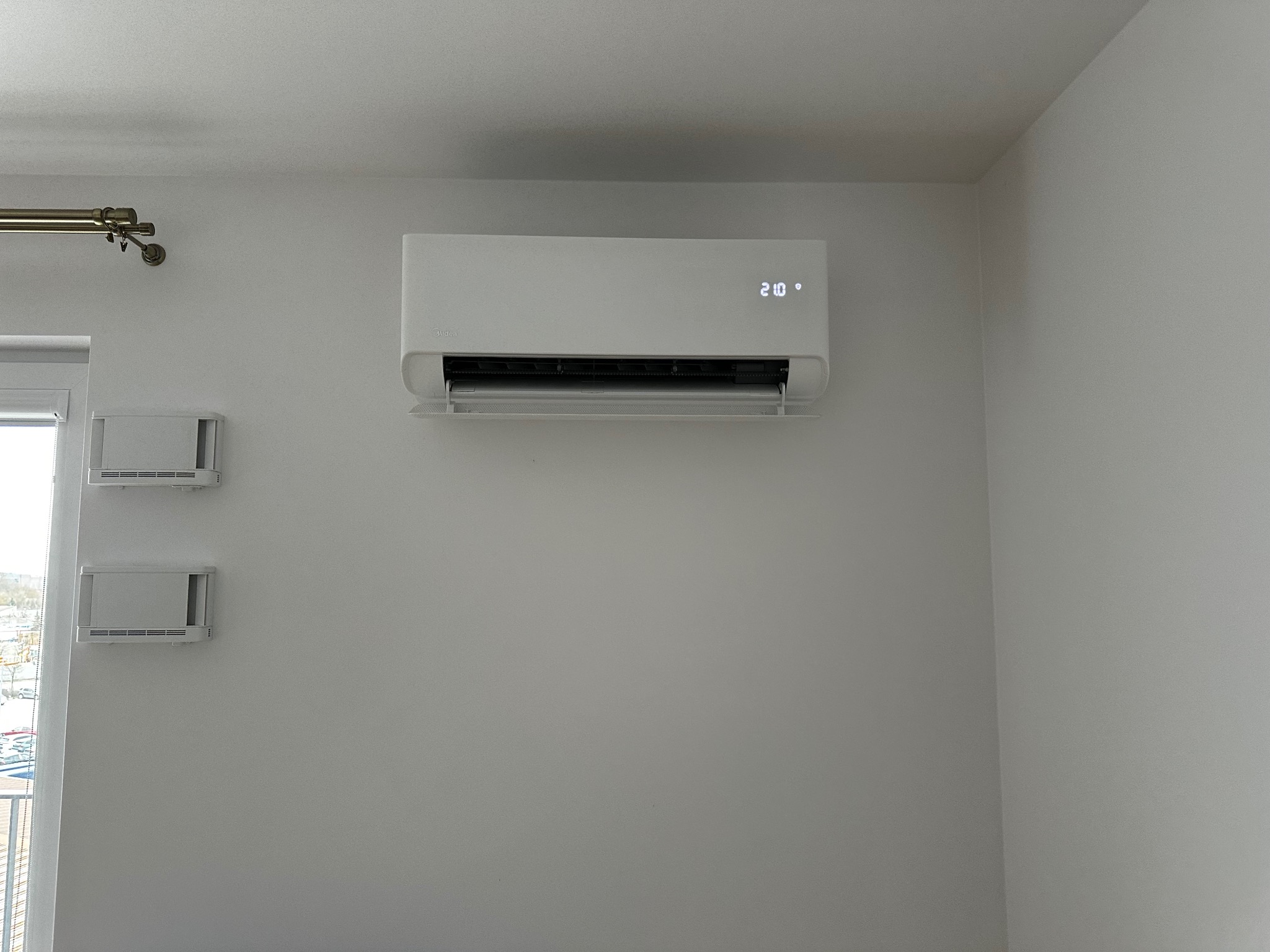 Biały klimatyzator Midea z wyświetlaczem 21 stopni Celsjusza zamontowany na ścianie w jasnym pomieszczeniu. Widoczne kratki wentylacyjne i karnisz.