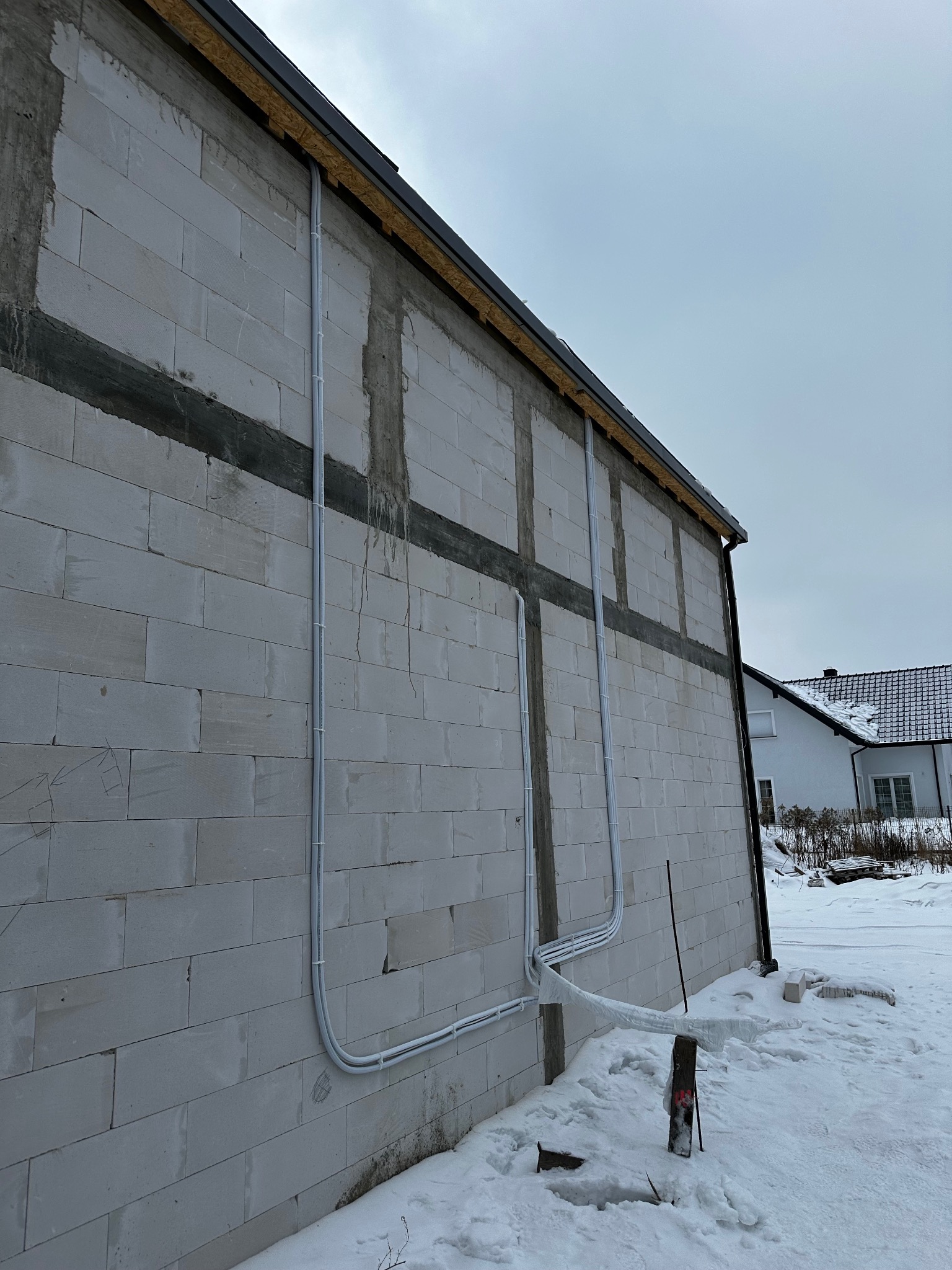Białe rury instalacyjne prowadzone po zewnętrznej ścianie budynku z betonu komórkowego w zimowej scenerii. Widoczne mocowania rur i fragment dachu.