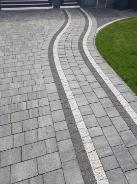 Ułożona z szarej kostki brukowej ścieżka o falującym kształcie, obramowana białymi i ciemnoszarymi kostkami, prowadząca do furtki i bramy wjazdowej, z fragmentem trawnika po prawej stronie.