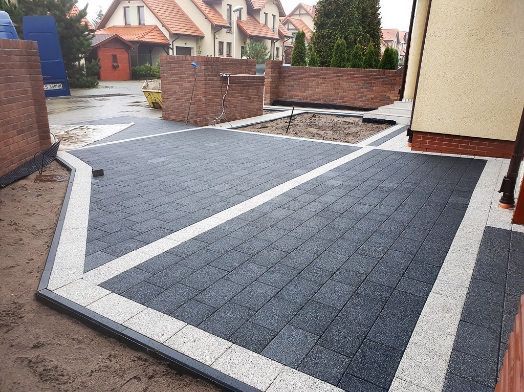 Ułożona ciemnoszara kostka brukowa z białymi pasami wyznaczającymi granice, otoczona piaskiem i elementami małej architektury z cegły klinkierowej, widoczne budynki mieszkalne w tle.