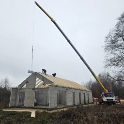 KAM-BUD Kamil Andrych - Montaż prefabrykowanego domu z użyciem dźwigu: robotnicy na dachu układają drewnianą konstrukcję więźby dachowej na betonowych ścianach, dźwig podnosi kolejny element, w tle szary krajobraz.