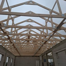 KAM-BUD Kamil Andrych - Wnętrze budynku w stanie surowym otwartym z widoczną drewnianą konstrukcją więźby dachowej, połączoną metalowymi płytkami, na tle pochmurnego nieba.