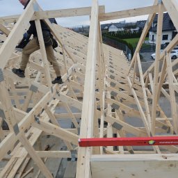 KAM-BUD Kamil Andrych - Konstrukcja drewniana więźby dachowej w Pyszkowie, widok z góry na pracującego dekarza z narzędziem, w tle zabudowa jednorodzinna.