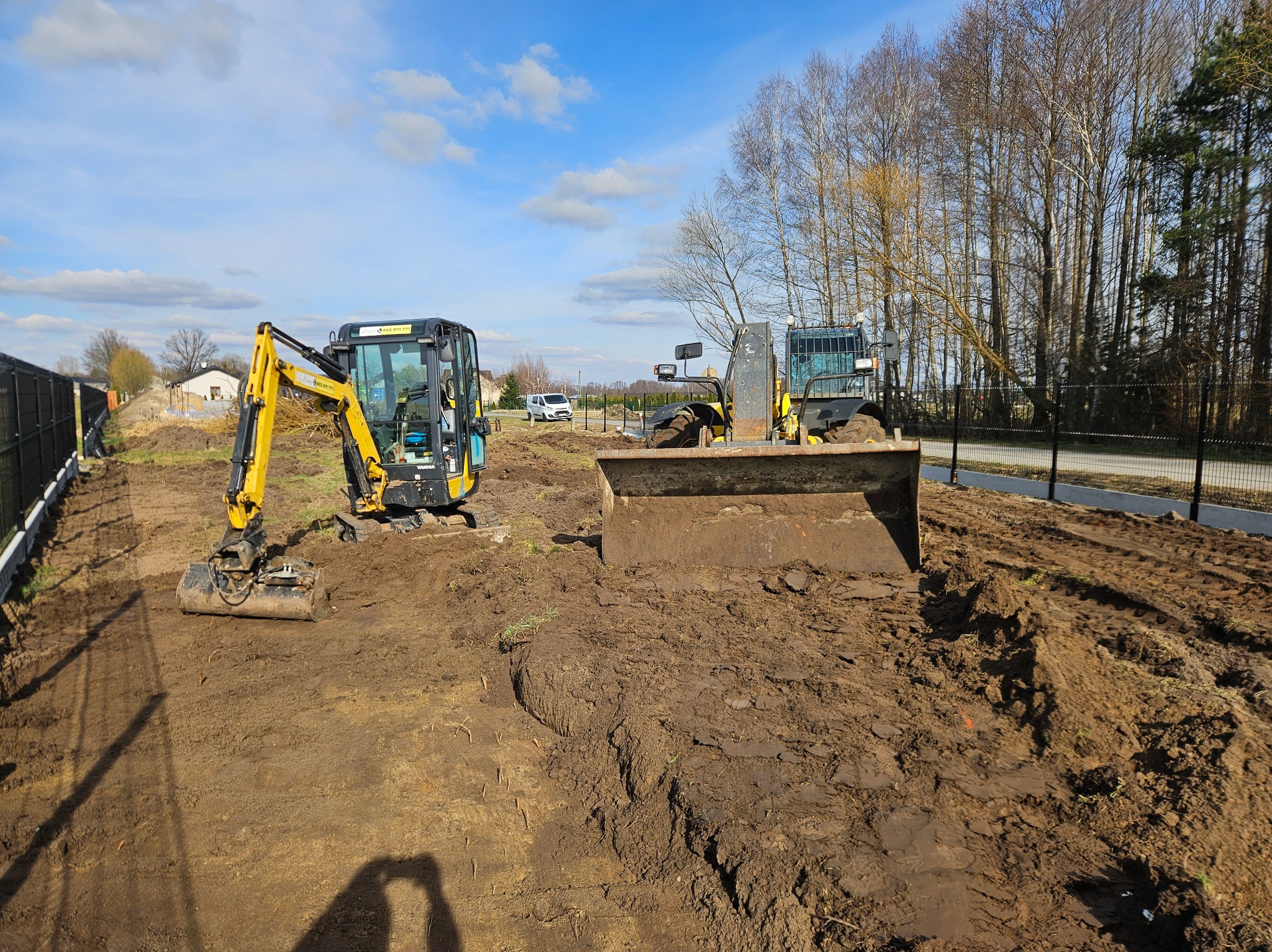 Dwa żółte pojazdy budowlane, w tym mała koparka i spychacz, na placu budowy z ziemią, w tle domy i drzewa pod błękitnym niebem.