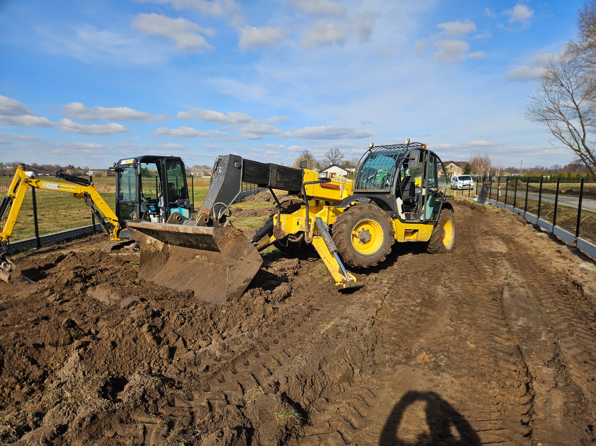 Żółta ładowarka teleskopowa i mała koparka na placu budowy, świeżo wykopana ziemia, w tle ogrodzenie panelowe i błękitne niebo z chmurami.