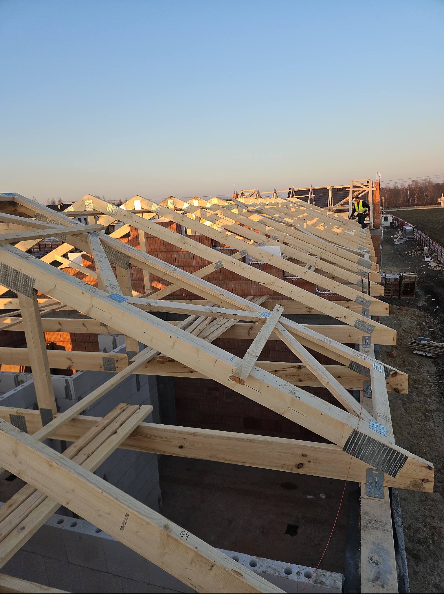 Konstrukcja drewniana więźby dachowej z widocznymi elementami złącznymi na tle muru z czerwonej cegły i błękitnego nieba o zachodzie słońca. W tle widoczna sylwetka pracownika w kamizelce odblaskowej.