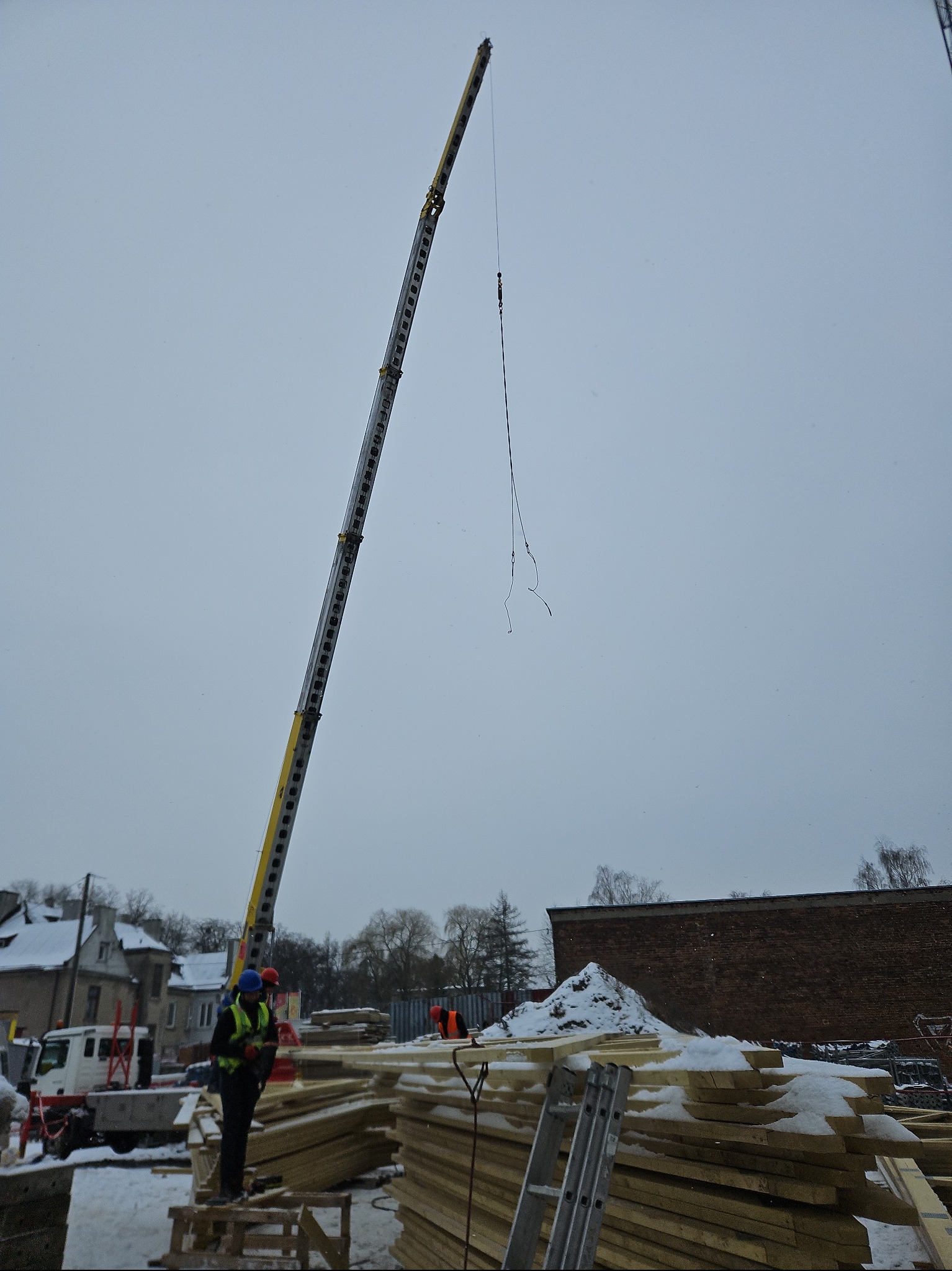 Praca dźwigu podczas montażu konstrukcji drewnianej dachu w zimowych warunkach, widoczne elementy więźby dachowej i pracownicy w odblaskowych kamizelkach.
