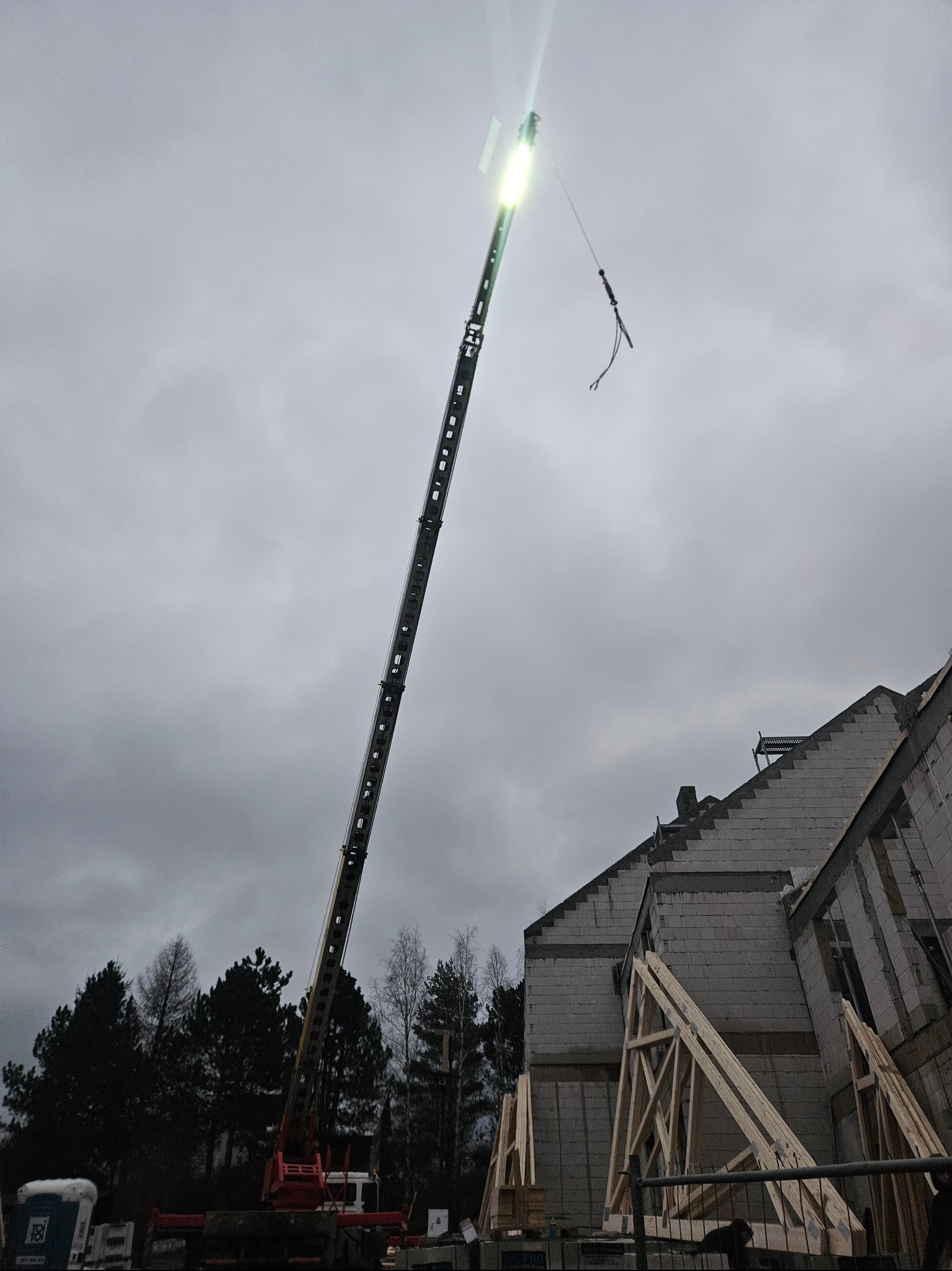Żuraw budowlany z podwieszonym elementem nad powstającą konstrukcją dachu domu jednorodzinnego, widoczne elementy więźby dachowej, pochmurne niebo w tle.