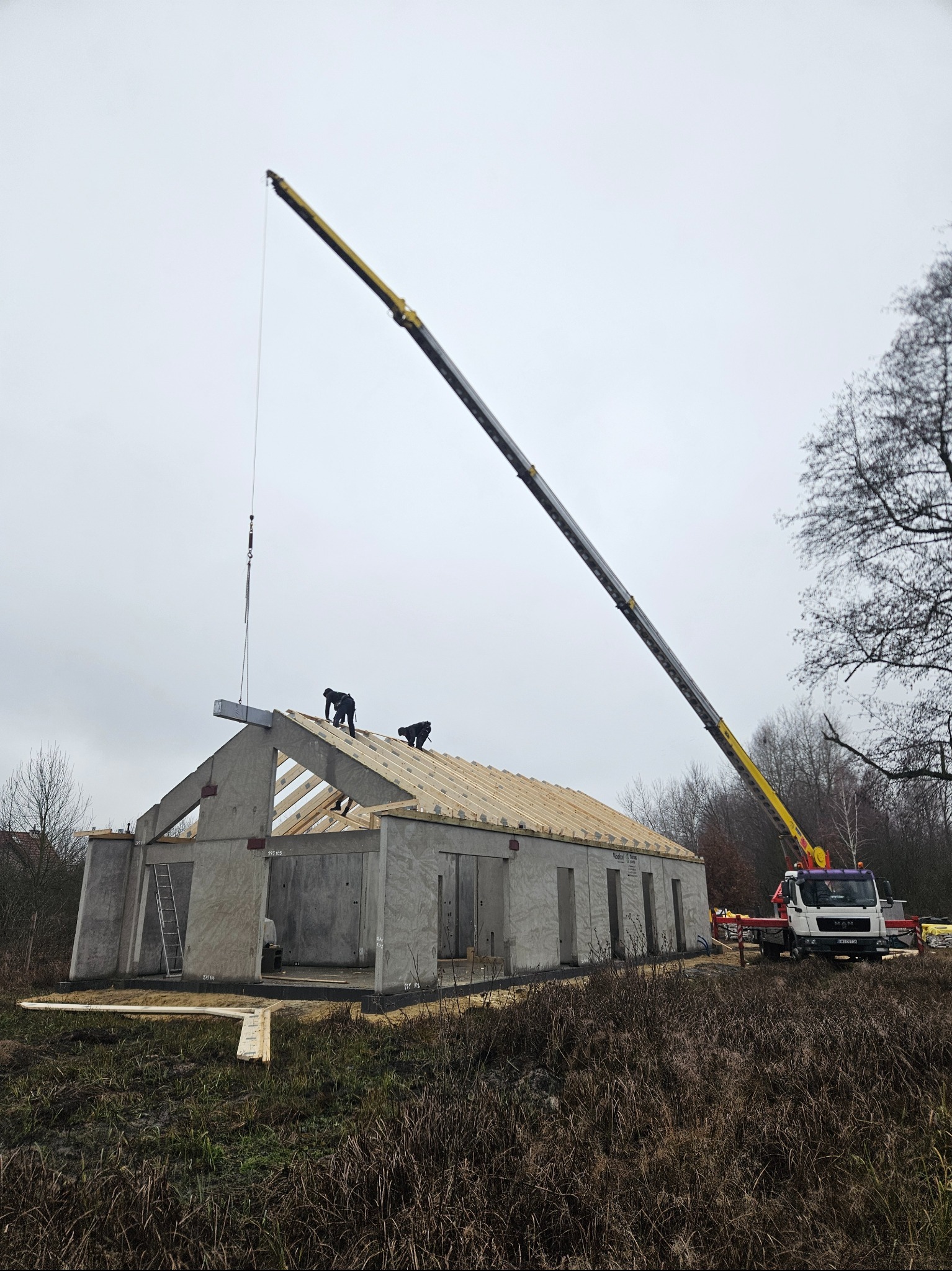 Montaż prefabrykowanego domu z użyciem dźwigu: robotnicy na dachu układają drewnianą konstrukcję więźby dachowej na betonowych ścianach, dźwig podnosi kolejny element, w tle szary krajobraz.