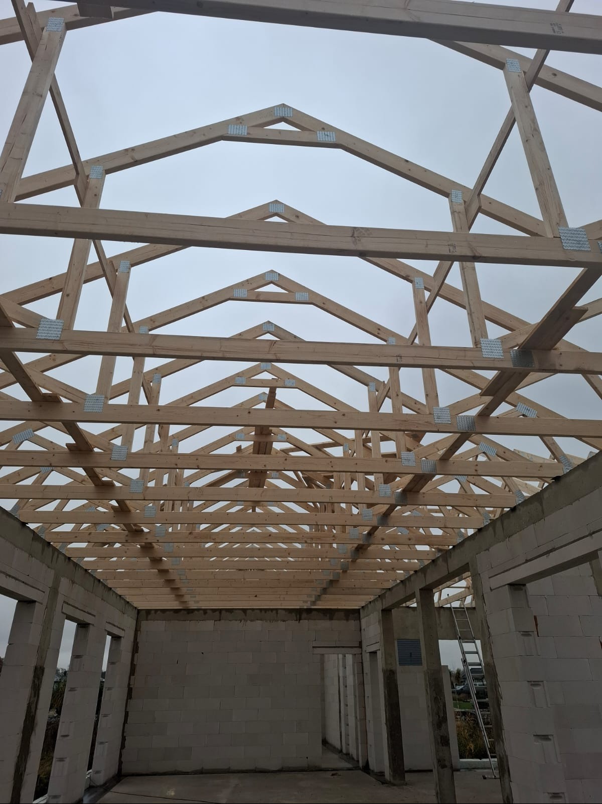 Wnętrze budynku w stanie surowym otwartym z widoczną drewnianą konstrukcją więźby dachowej, połączoną metalowymi płytkami, na tle pochmurnego nieba.