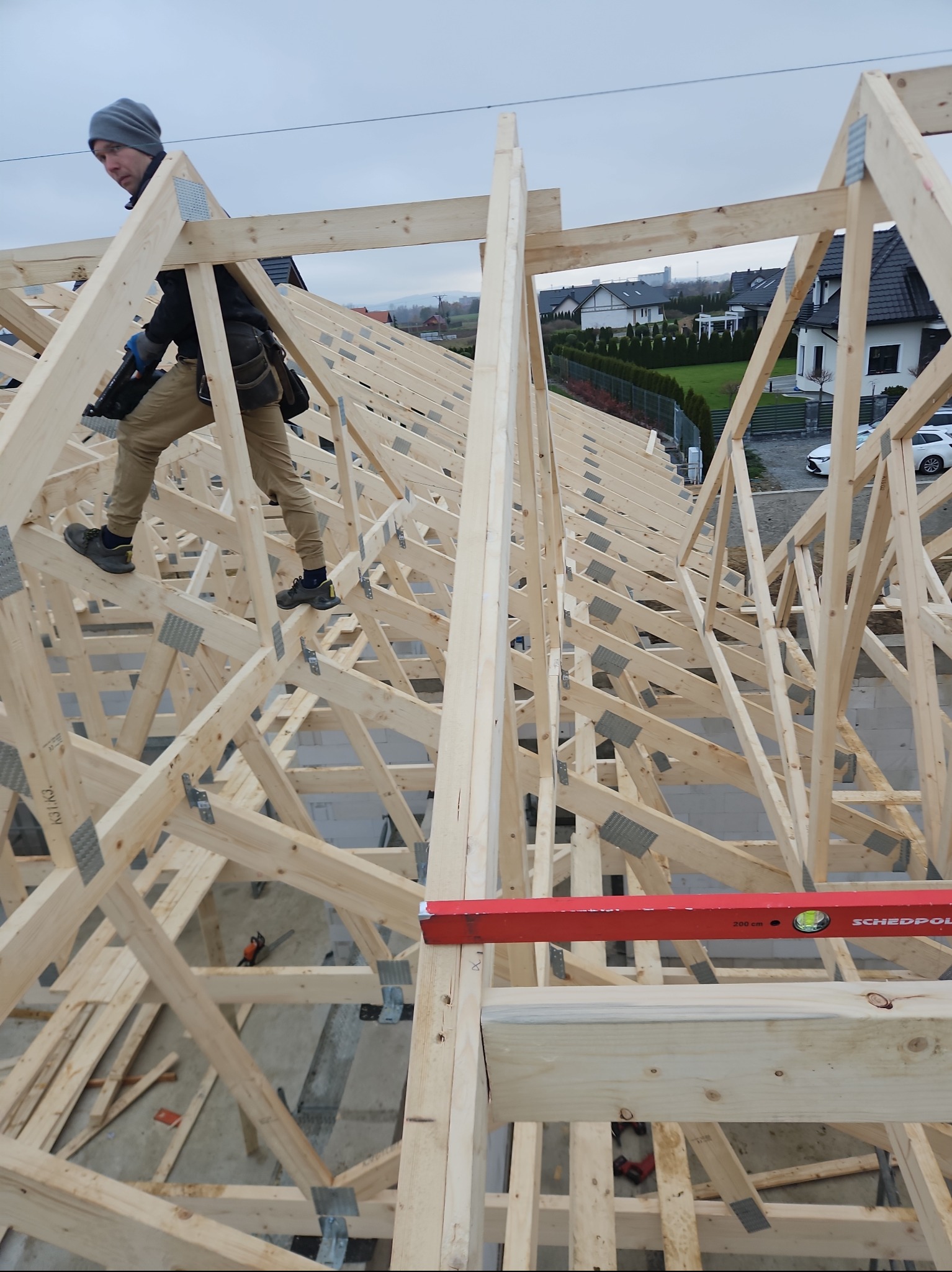 Konstrukcja drewniana więźby dachowej w Pyszkowie, widok z góry na pracującego dekarza z narzędziem, w tle zabudowa jednorodzinna.