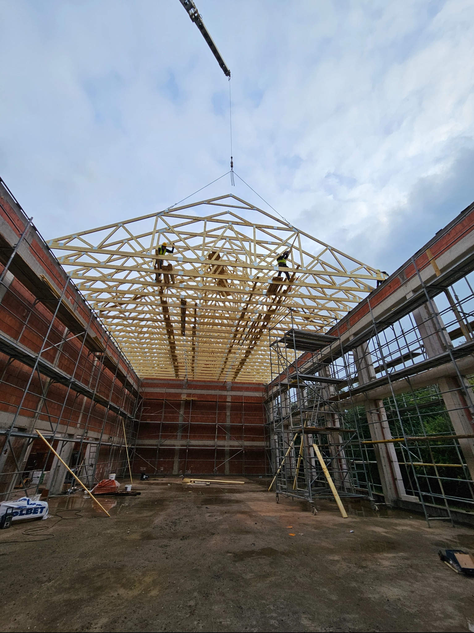 Montaż drewnianej więźby dachowej wewnątrz budynku w trakcie budowy, widoczni dwaj pracownicy na rusztowaniach, dźwig podnosi elementy konstrukcji.