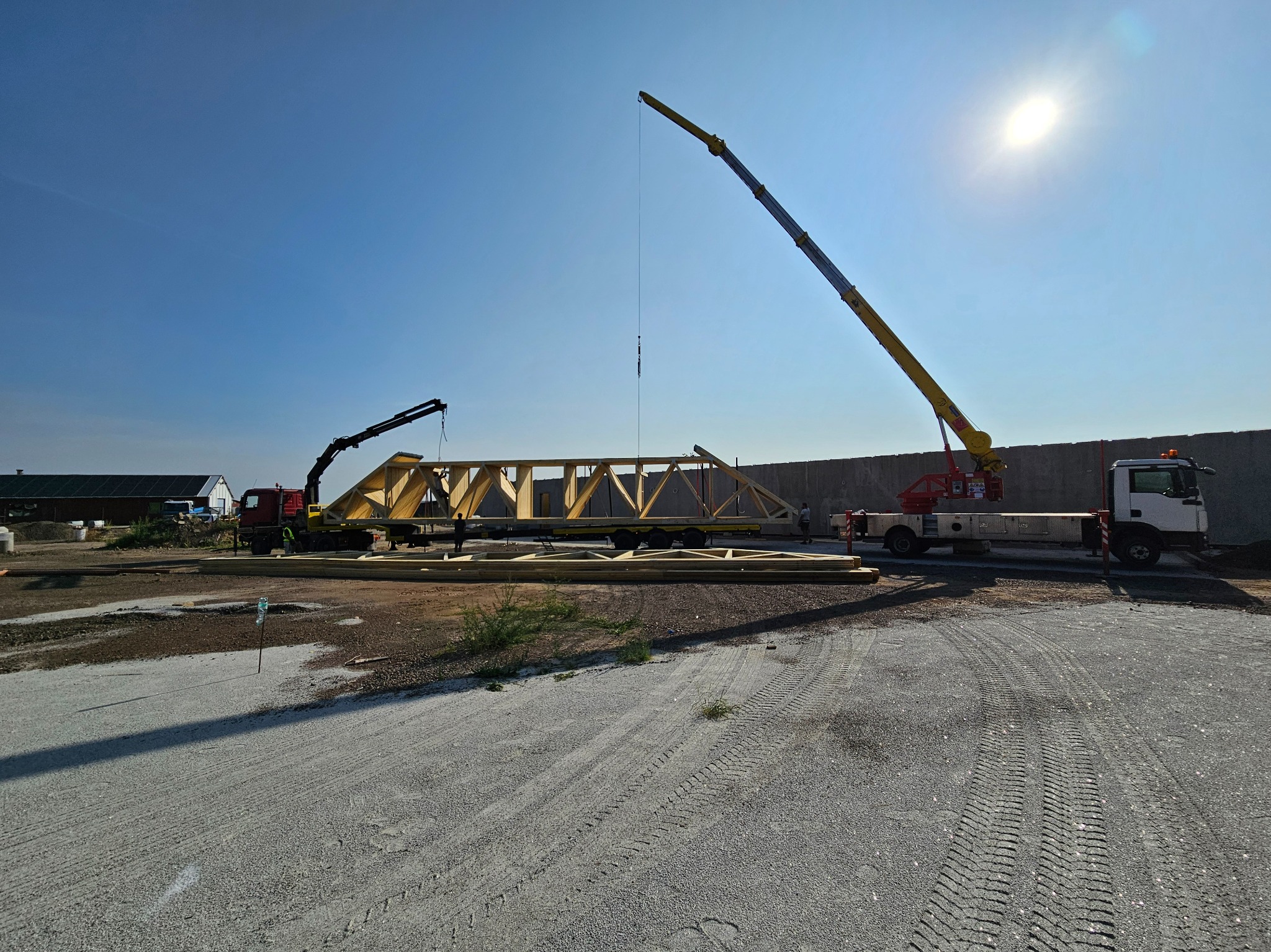 Dwa dźwigi podnoszą drewnianą konstrukcję więźby dachowej na placu budowy w słoneczny dzień.