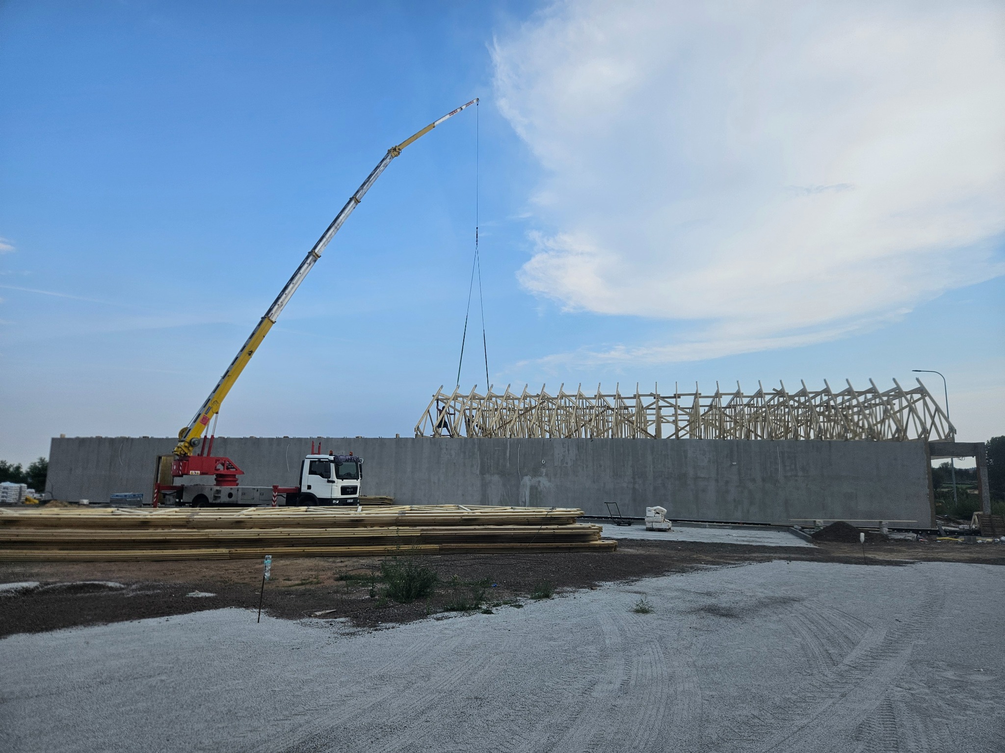 Żuraw podnosi element drewnianej konstrukcji dachu na placu budowy, widoczne stosy drewna i betonowa ściana.