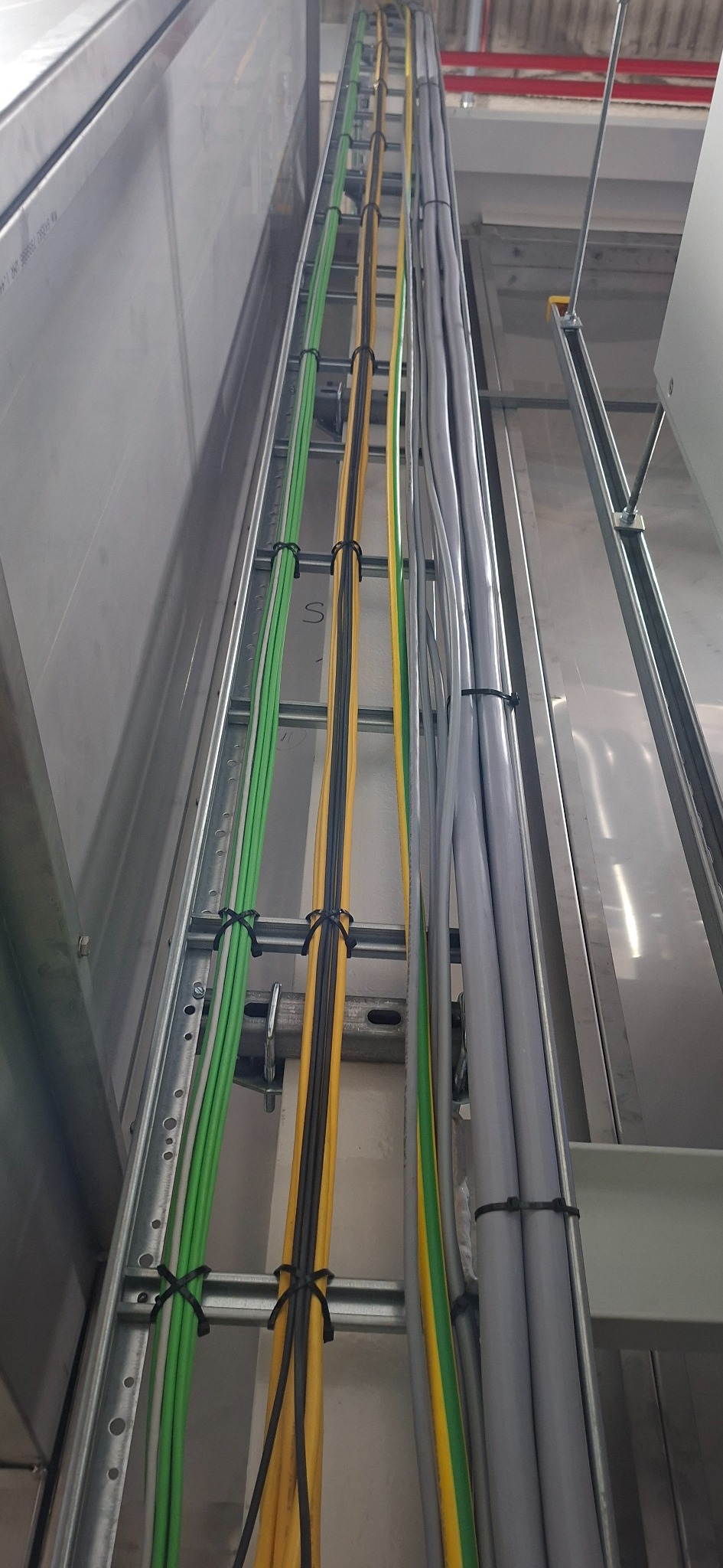 Rząd przewodów elektrycznych w kolorach: zielonym, żółtym, czarnym i szarym, ułożonych w metalowej drabince, zamocowanych czarnymi opaskami zaciskowymi.