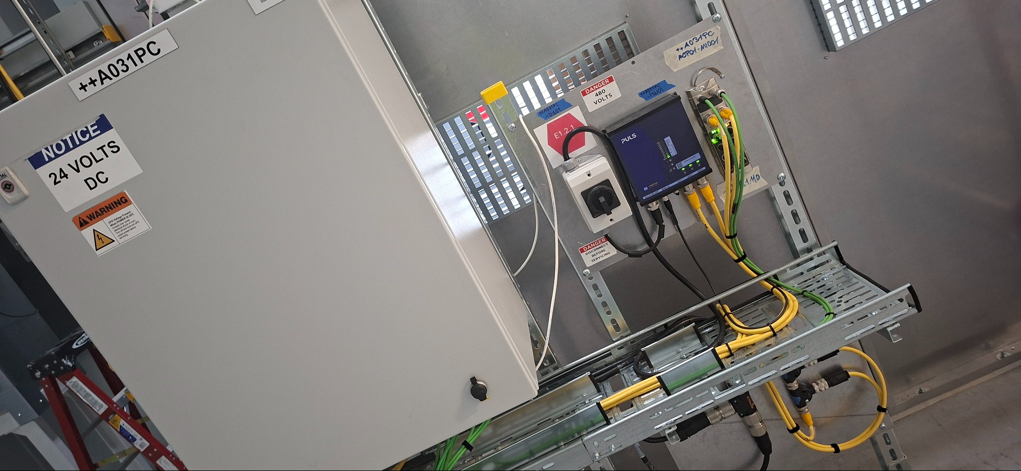 Szara szafa sterownicza z okablowaniem, etykietami ostrzegawczymi i urządzeniem Puls. Widoczne kable w żółtej i zielonej izolacji, drabinka kablowa i fragment drabiny.