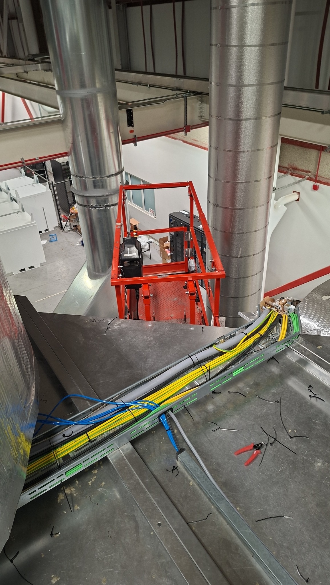Instalacja kabli w korycie na wysokości, widoczna z góry. W tle podnośnik koszowy i elementy wentylacji. Przewody w kolorach: żółtym, niebieskim i zielonym.