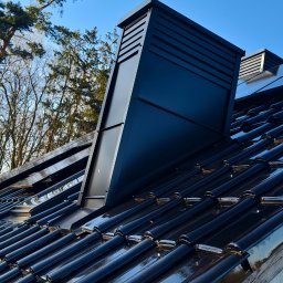 PASCALE SPÓŁKA Z OGRANICZONĄ ODPOWIEDZIALNOŚCIĄ - Błyszczący, czarny dach z ceramicznej dachówki karpiówki i nowoczesnym, czarnym kominem wentylacyjnym na tle drzew i błękitnego nieba.