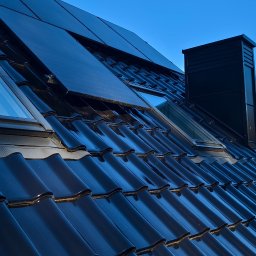 PASCALE SPÓŁKA Z OGRANICZONĄ ODPOWIEDZIALNOŚCIĄ - Błyszczący, czarny dach z dachówki ceramicznej z oknami dachowymi, kominem i panelami solarnymi, widok z dołu pod ostrym kątem na tle błękitnego nieba.