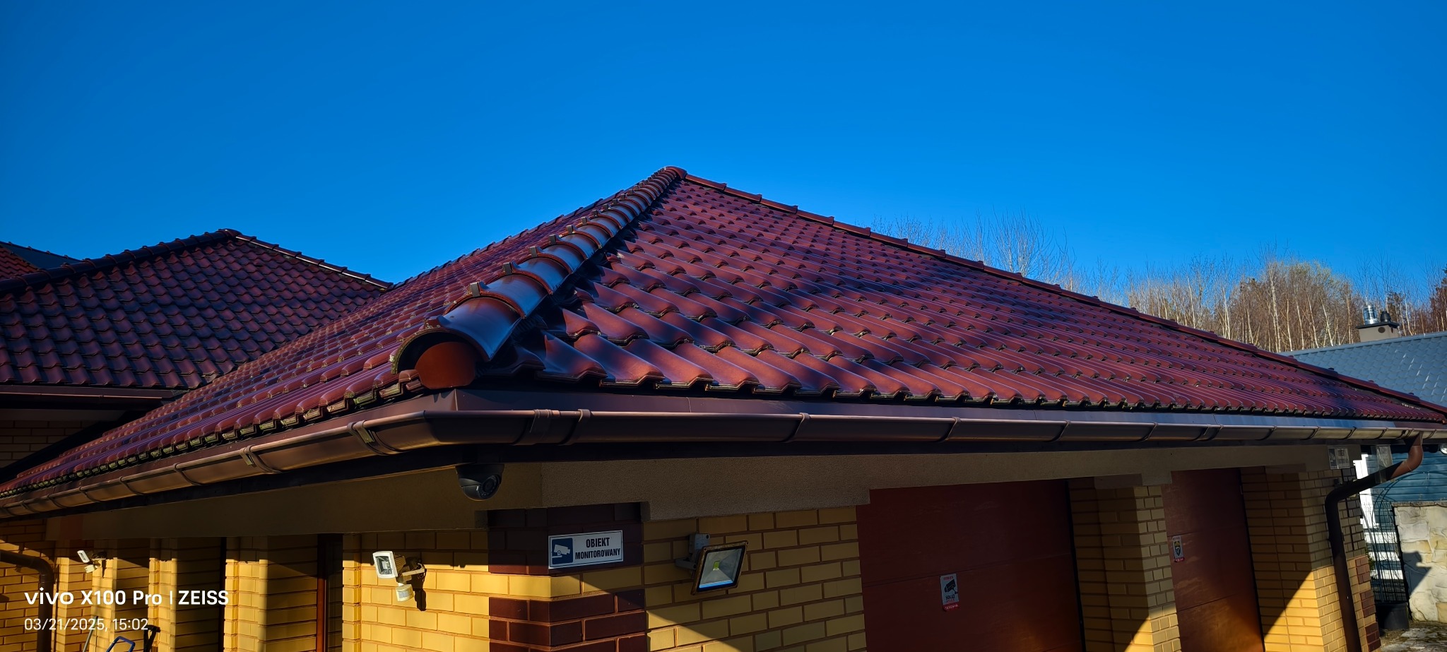 Nowy, lśniący dach z czerwonej dachówki ceramicznej, widoczny z dołu pod ostrym kątem, z zamontowanymi rynnami w kolorze brązowym, na tle błękitnego nieba i fragmentu ściany budynku z cegły...