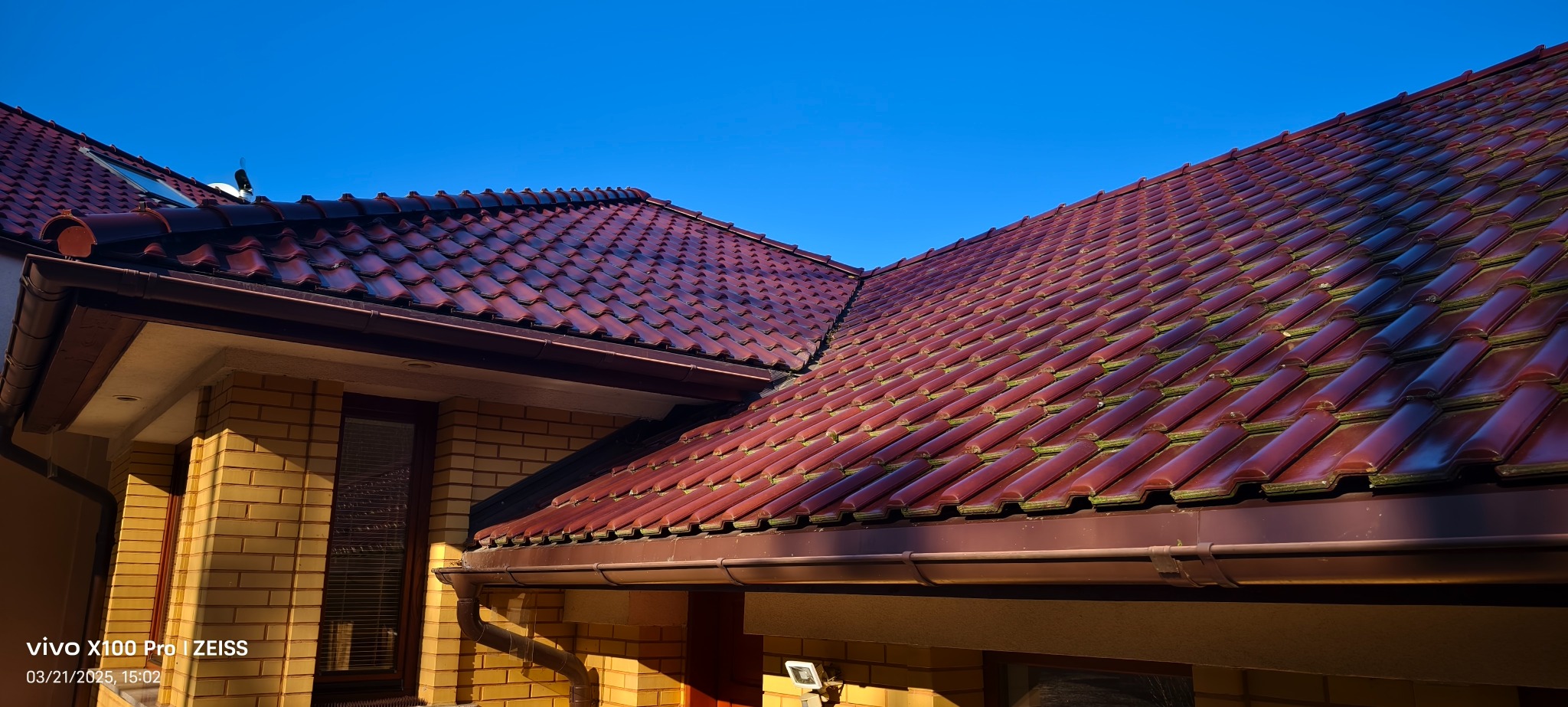 Czerwona dachówka ceramiczna na skośnym dachu domu jednorodzinnego, widoczne rynny w kolorze brązowym i fragment elewacji z cegły klinkierowej w odcieniach żółci.