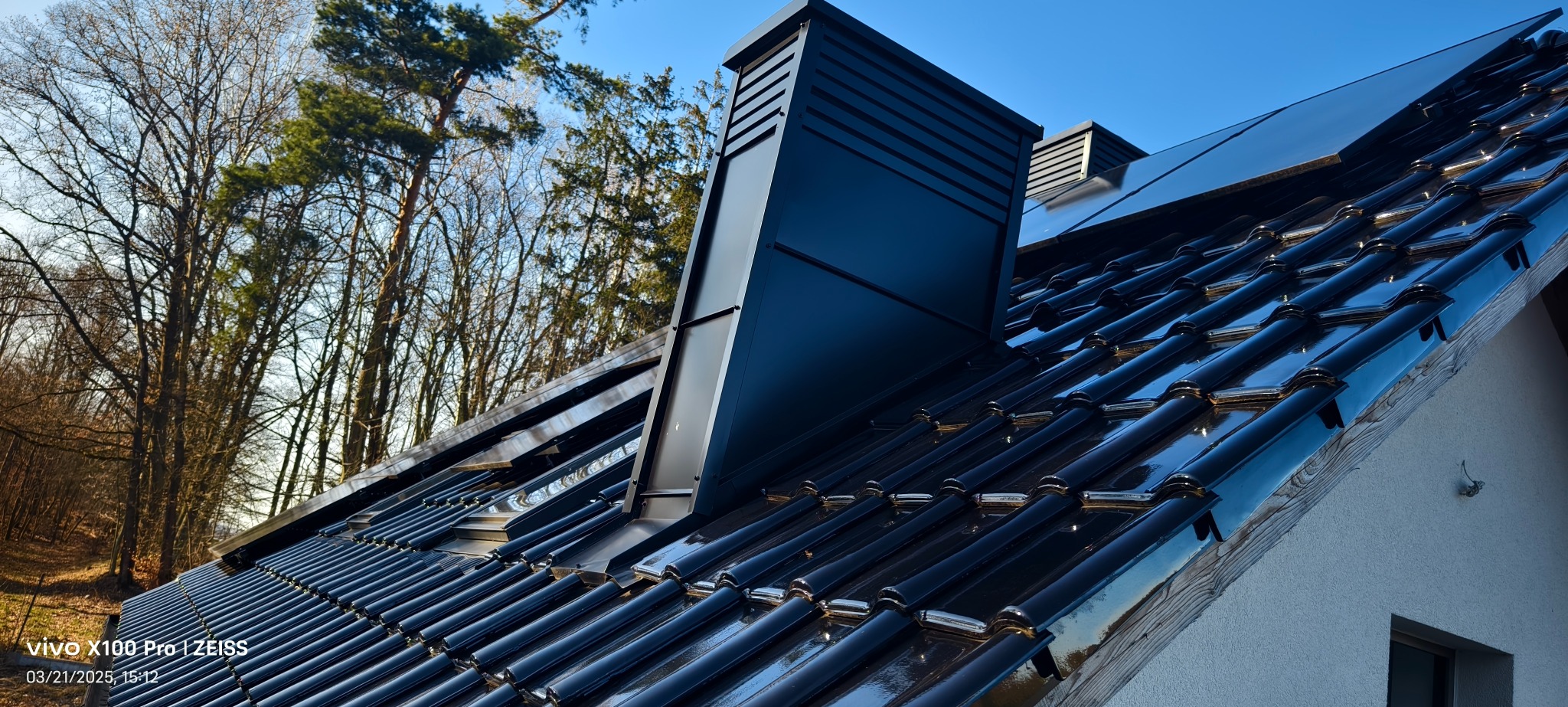 Błyszczący, czarny dach z ceramicznej dachówki karpiówki i nowoczesnym, czarnym kominem wentylacyjnym na tle drzew i błękitnego nieba.