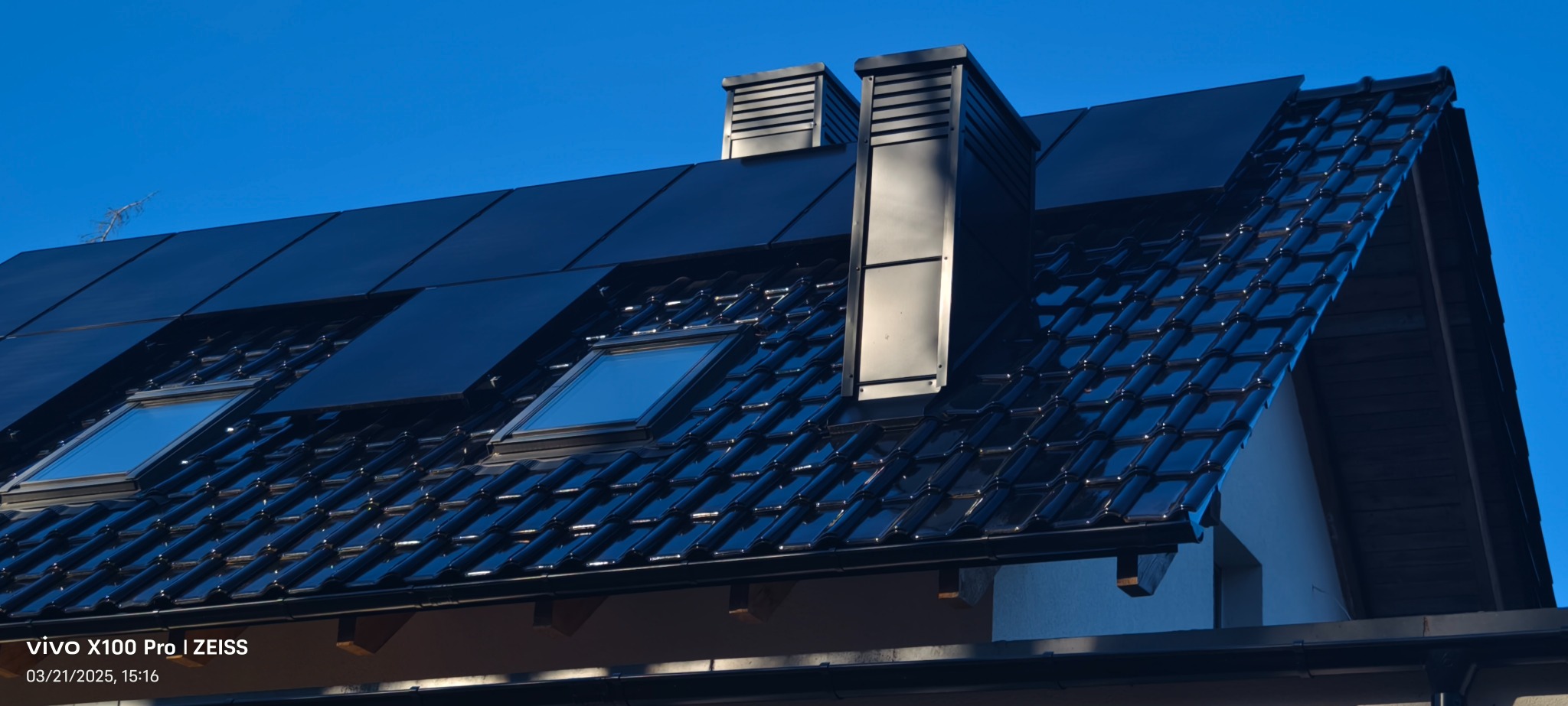 Czarny dach z dachówki ceramicznej z oknami dachowymi i kominami wentylacyjnymi na tle błękitnego nieba.